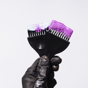 Gloved hand holding two hair color brushes, one with white dye and the other with purple color, against a plain white background. 