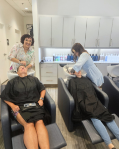 Two women sitting in salon chairs get their hair washed by two stylists in a modern, well-lit hair salon. Bottles of hair products are visible on shelves in the background. - Salon Jaylee | The Villages, FL
