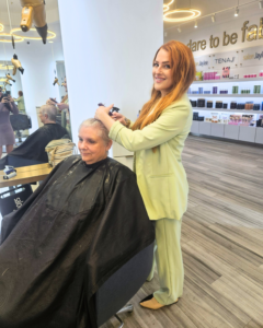 A stylist in a light green suit shaves the head of a seated client in a modern hair salon with shelves of products in the background. - Salon Jaylee | The Villages, FL